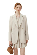 A woman models a light beige double-breasted blazer over a matching beige pleated mini dress, holding a small brown leather handbag against a plain off-white studio background.