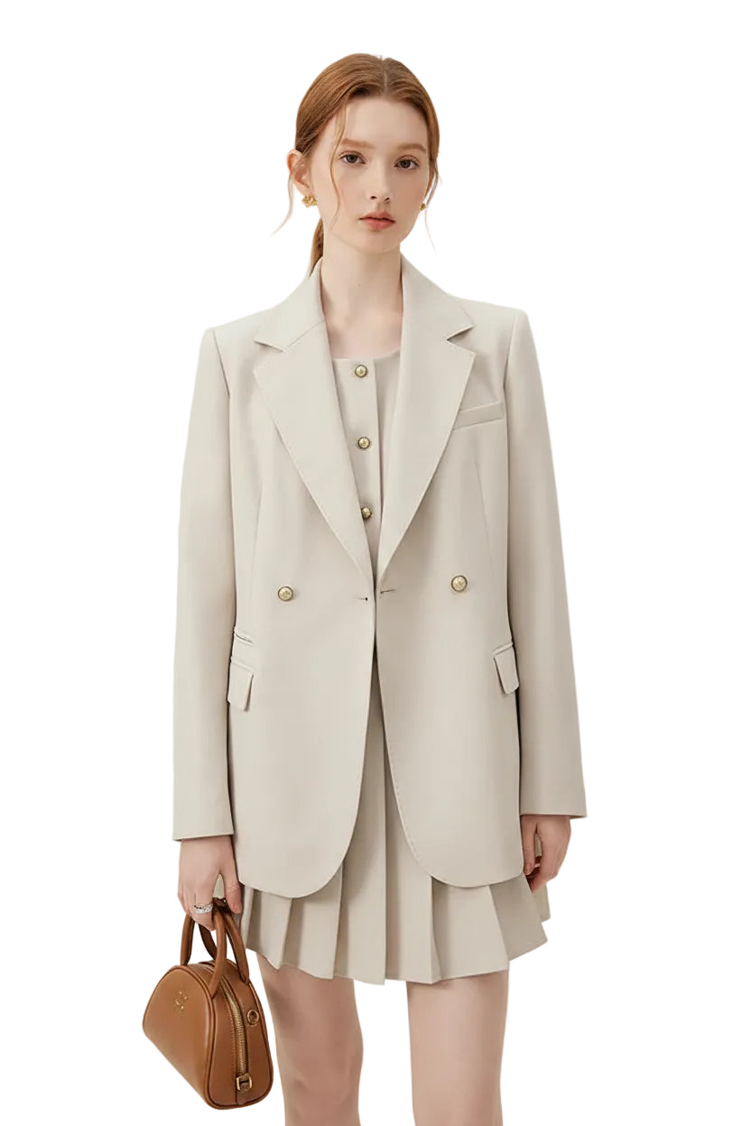 A woman models a light beige double-breasted blazer over a matching beige pleated mini dress, holding a small brown leather handbag against a plain off-white studio background.