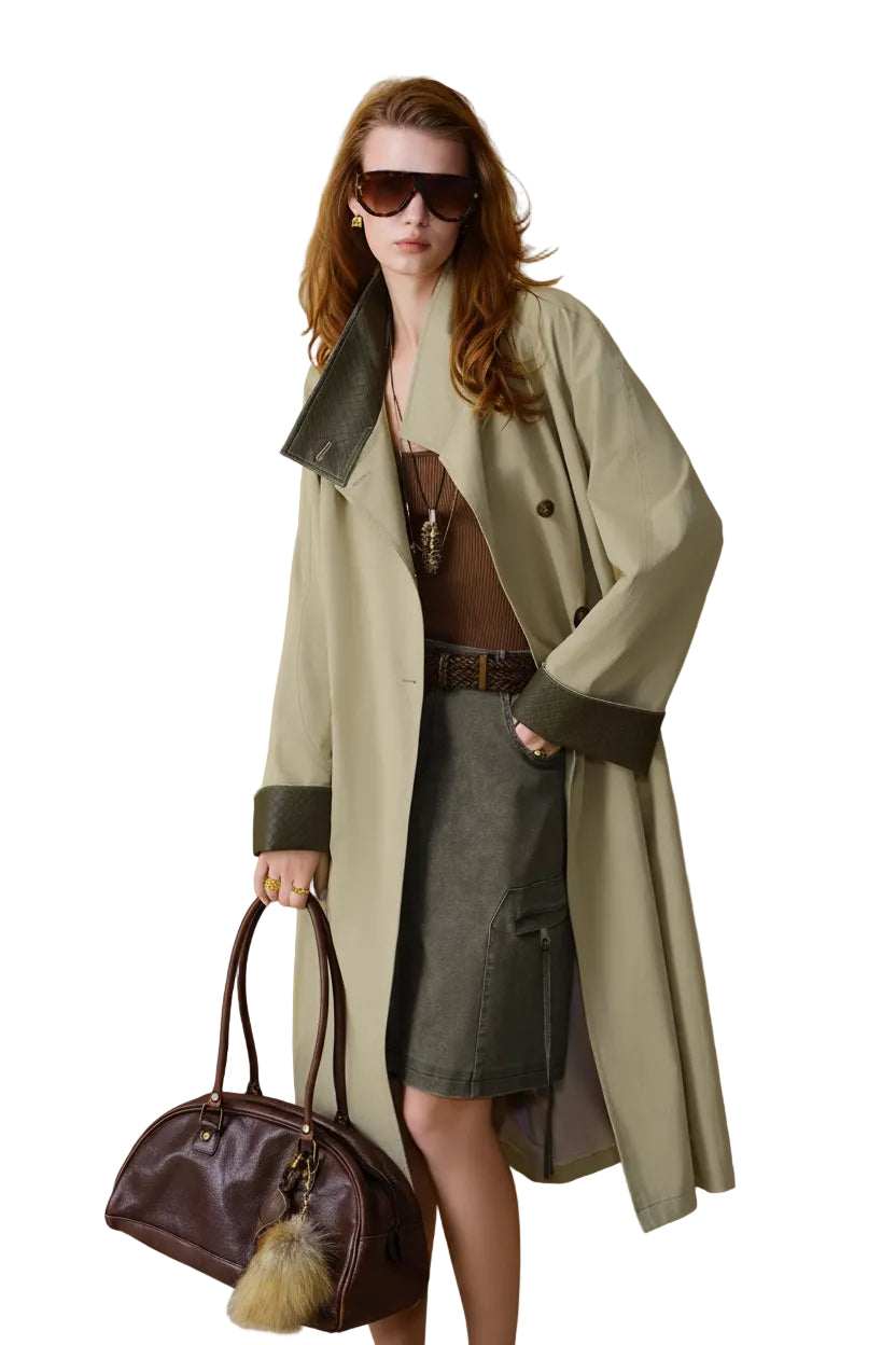 A woman models a long beige trench coat with dark green leather trim, styled with a brown top, a gray skirt, sunglasses, and a dark brown handbag against a textured gold background.