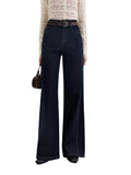 A model is shown from the waist down wearing dark navy wide-leg denim jeans, a brown belt, a cream patterned long-sleeve top, and black shoes, holding a small studded brown handbag. The brand logo 'fsle' is visible in the top left corner.